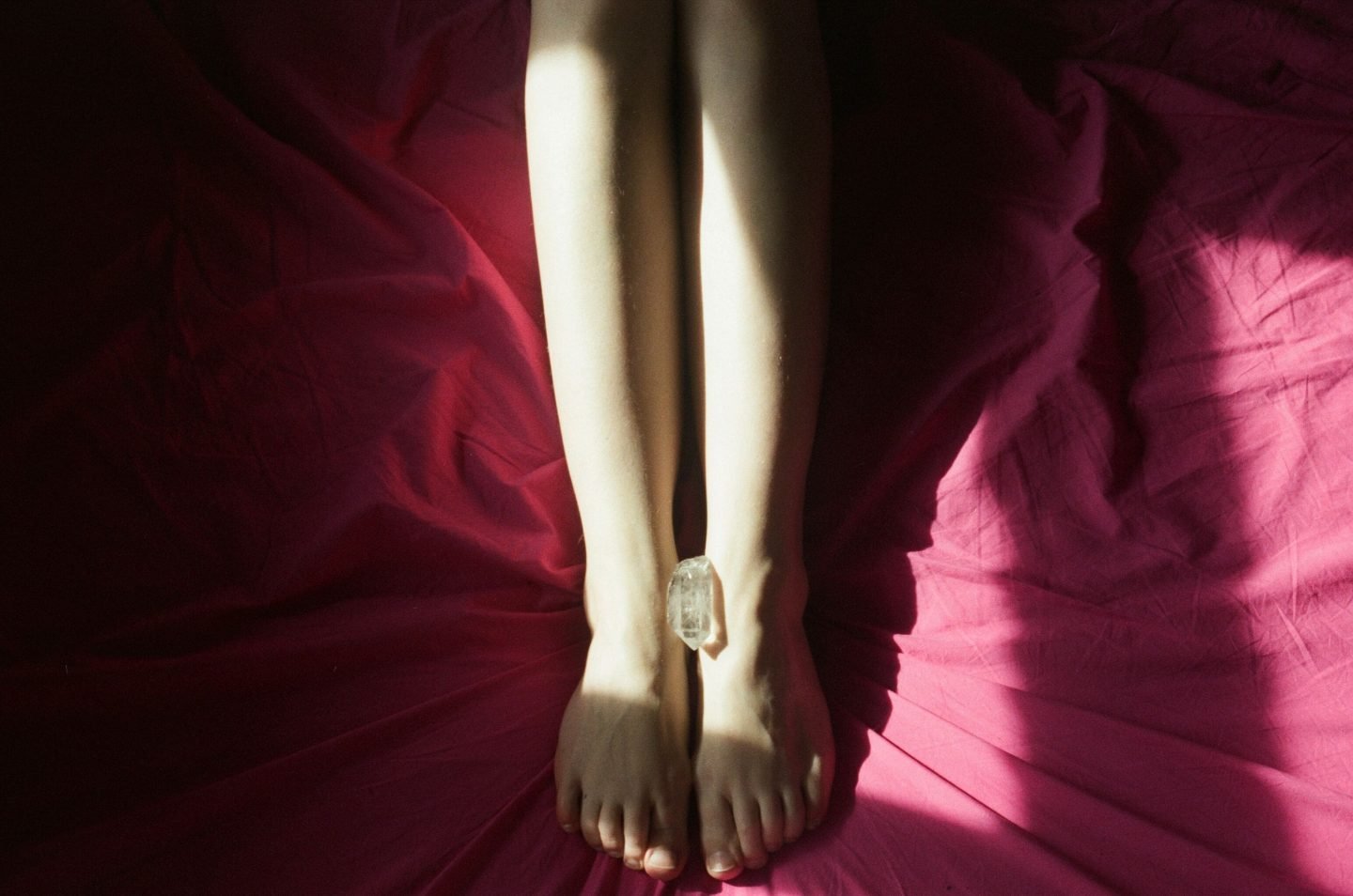 alt=“a photoof a piece of crystal is placed between a woman’s feet who is in bed on red bedsheets from Lina Scheynius photo - essay for Tabayer jewelry ”