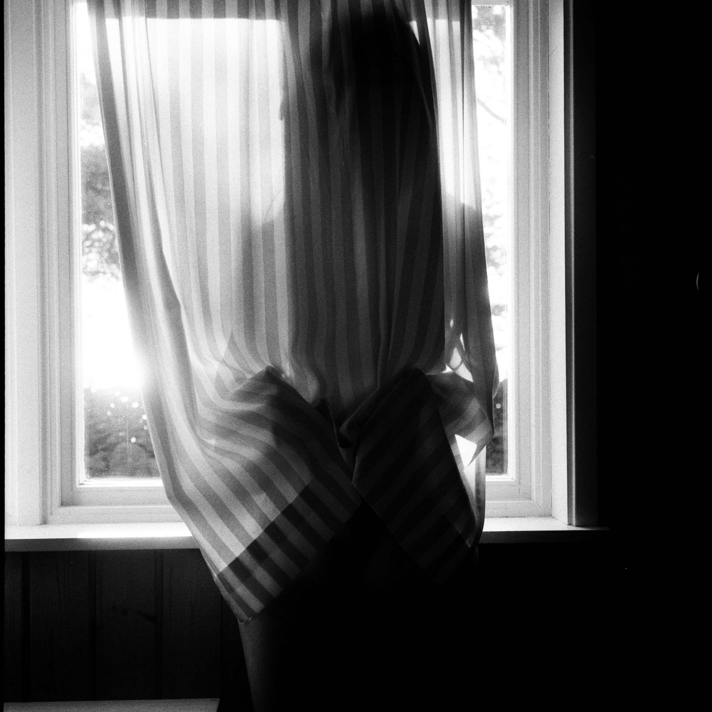 alt=“a black and white photo of a woman standing by the window behind a curtain from Lina Scheynius photo - essay for Tabayer jewelry ”