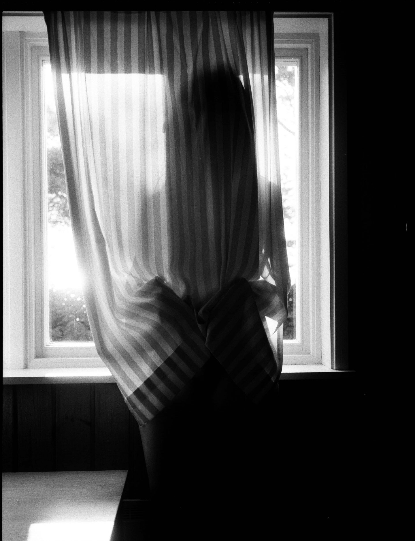 alt=“a black and white photo of a woman standing by the window behind a curtain from Lina Scheynius photo - essay for Tabayer jewelry ”